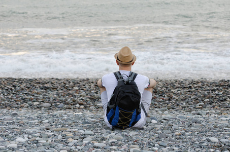 年轻男子戴着帽子和背包里坐在沙滩上的大海背景上。查看从背面