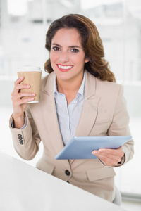 一位兴高采烈的女商人拿着一次性杯子在办公桌上使用数字平板电脑