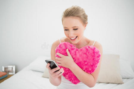 自然微笑的金发女郎拿着心形枕头和智能手机