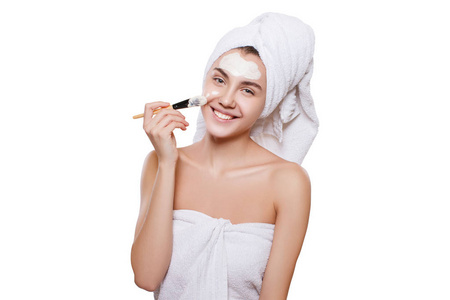 年轻漂亮的女孩用白色的毛巾在她的头上, 她的脸上有一个白色的保湿面具与刷子。粘土面膜, 化妆品, 治疗泥浆