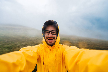 微笑和大笑旅行者人在黄色的雨衣，在 stromy 天气雨云山脉眼镜的自拍照肖像