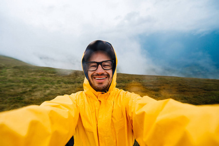 微笑和大笑旅行者人在黄色的雨衣，在 stromy 天气雨云山脉眼镜的自拍照肖像