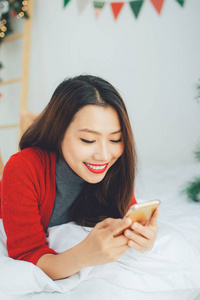 亚洲漂亮的女人躺在圣诞树附近与智能手机