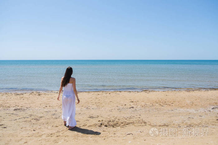 年轻的黑发女人,在夏天白色礼服站在海滩上,望海