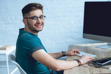 英俊的年轻人在眼镜工作与台式计算机和微笑在照相机