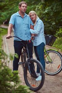 有吸引力的一对金发女郎和身着便装的男士, 在公园附近的自行车上拥抱