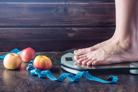 女性脚站立在电子秤为重量控制与测量磁带和苹果在黑暗的背景。运动训练饮食和减肥的概念
