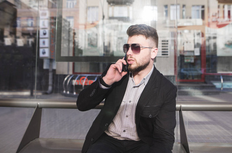 一个商人坐在公共汽车站, 通过电话交谈。一个穿着服装的时髦男士通过电话在公共交通站的长凳上讲话。
