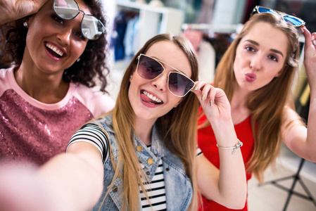 微笑的女朋友开心的时尚太阳镜时间以自拍照用手机做在服装店购物时