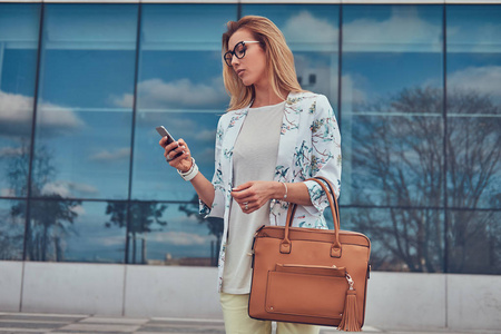 时尚的女性博客在时尚的衣服和眼镜与手提包, 持有智能手机, 站在摩天大楼上