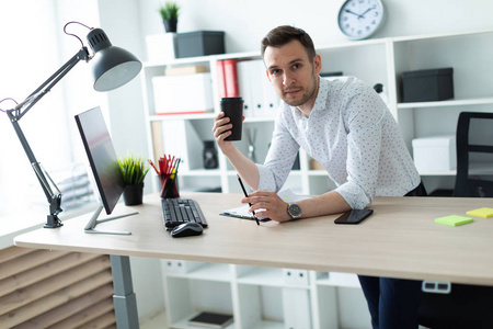 一个年轻人站在办公室的桌子旁, 拿着一支铅笔和一杯咖啡。一个年轻人工作的文件和计算机
