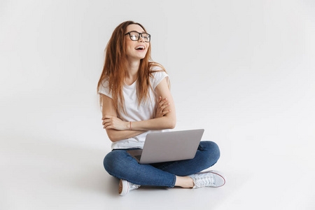 穿着 t恤和眼镜的开朗女人坐在地板上用笔记本电脑, 同时手持交叉双臂, 仰望灰色背景