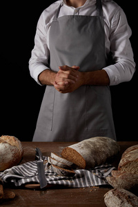 用刀麻布和面包站在桌子旁边的围裙上的男面包师的裁剪镜头