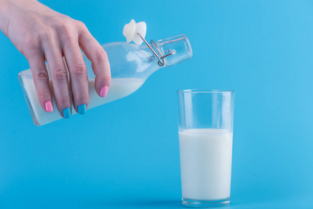 一个女人的手把新鲜的牛奶从玻璃瓶倒入蓝色背景的玻璃杯中。多彩简约。健康乳制品与钙的概念