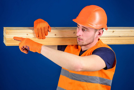 木匠, 木工, 强的建设者在繁忙的面孔运载木横梁在肩膀上。男子头盔, 硬帽和防护手套指向方向, 蓝色背景。木质材料概念