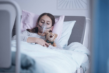 戴着氧气面罩的生病的女孩睡在医院的床上, 泰迪熊