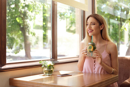 一个年轻的女人与梅森瓶美味的天然柠檬水在餐桌上的咖啡馆。排毒饮料