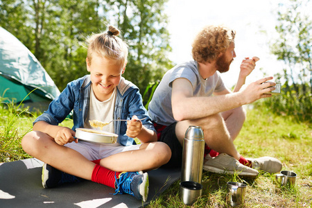 愉快的男孩吃粥与他的父亲坐在他旁边在地面在他们的旅行期间