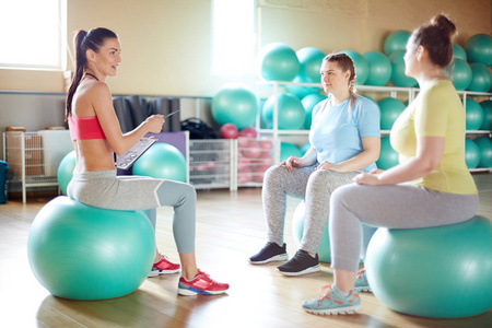 年轻的健身教练和她的小组坐在绿色 fitballs 在健身房和讨论他们要做的运动