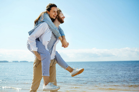 笑年轻的女人拥抱男人, 而坐在他的背部和享受夏天的海边