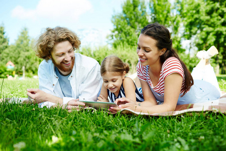 年轻夫妇和他们的女儿在绿色草坪上放松, 看着触摸板上的好奇视频