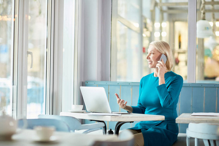 自信成熟的女商人在智能 casualwear 坐在咖啡桌前的笔记本电脑和客户交谈的智能手机