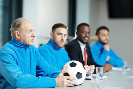 成熟的足球教练与足球和他的团队坐在一排在体育会议上