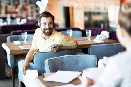 愉快的商人与智能手机看他的同事, 而坐在软扶手椅在咖啡馆