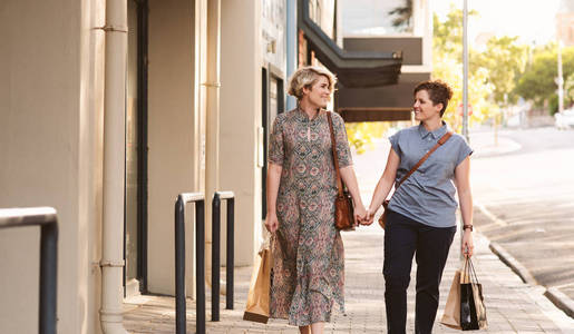 微笑的年轻女同志夫妇手牵手走在城市的街道上背着购物袋, 同时享受一天一起出去
