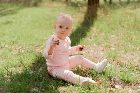 婴儿坐在草地上, 玩着两个冷杉锥, 她穿着粉红色的上衣对着绿草
