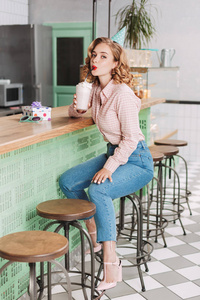 美丽的女士在生日帽坐在酒吧柜台和喝奶昔, 而朦胧地看着在咖啡馆的相机