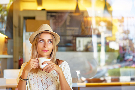 一个漂亮的年轻女子在帽子捧杯饮料, 而坐在咖啡馆