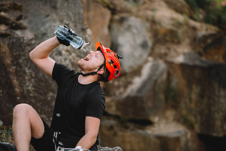 英俊年轻的旅行者在头盔饮用水从塑料瓶在岩石前面