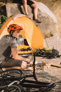 运动的年轻自行车旅行者坐在岩石上露营帐篷和模糊的朋友坐在背景上