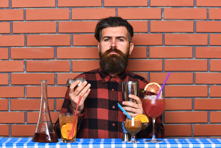 酒精饮料概念。酒保用长胡子和胡子和时髦的头发在严密的面孔举行的振动筛, 做酒精鸡尾酒。墙上穿格子衫的人准备饮料