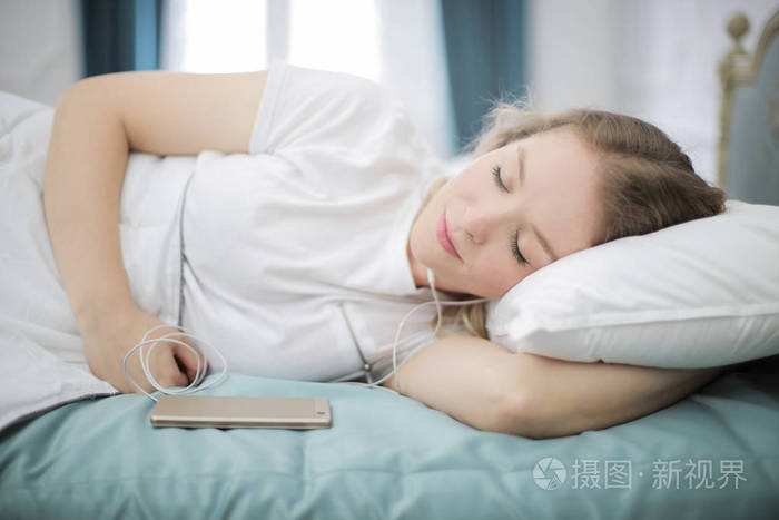 年轻的金发女人躺在她的床上, 放松, 而听音乐在她的智能手机