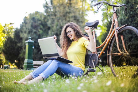 年轻卷曲头发的妇女坐在公园与她的笔记本电脑和自行车, 感到非常兴奋