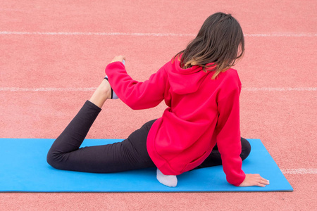 一个穿着红色 t恤和黑色裤子的女孩进行健身锻炼。露天的蓝色健身房垫。后退视图。在一个带粉红色跑步机的体育场内伸展训练