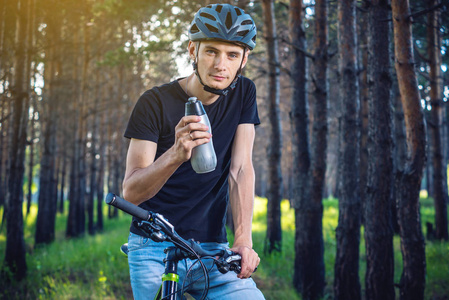 骑在头盔里的骑自行车的骑车人, 在树间的公园里坐着喝水。积极健康的生活方式概念
