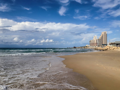 一个孤独的浪漫海滩的景色, 在沙滩上翻滚的波浪和高高的建筑物在蓝天下白云下的地平线上。照片是在以色列海法公共海滩拍摄的。