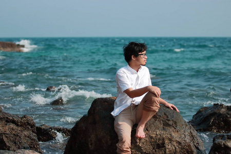 在海边的岩石上悠闲地坐在白色休闲的亚洲人。假期暑期概念