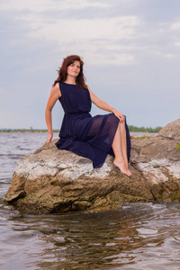 穿着黑色长裙的年轻漂亮女孩坐在河边的岩石上