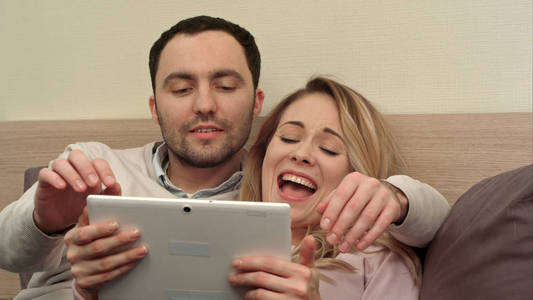 年轻的夫妇在床上使用数字平板电脑，微笑和大笑