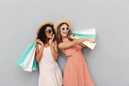 两个穿着夏季服装的快乐年轻女性的肖像手持购物袋, 指向在灰色背景下隔离的复制空间