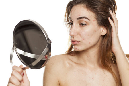 有问题的皮肤和没有化妆的年轻妇女看着自己在镜子上的白色背景