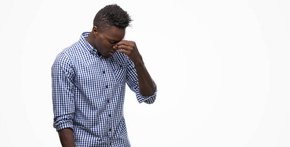 年轻的非洲裔美国人穿着蓝色衬衣疲倦地擦鼻子和眼睛感到疲倦和头疼。压力和挫折概念