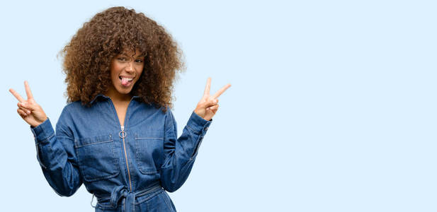 非洲裔美国妇女穿着蓝色的连身裤看着相机显示舌头和取得胜利标志用手指