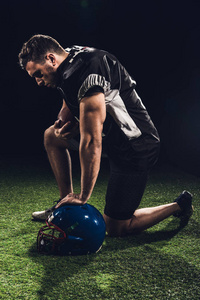 年轻严肃的美国足球运动员站在草地上的一个膝盖上的黑色头盔