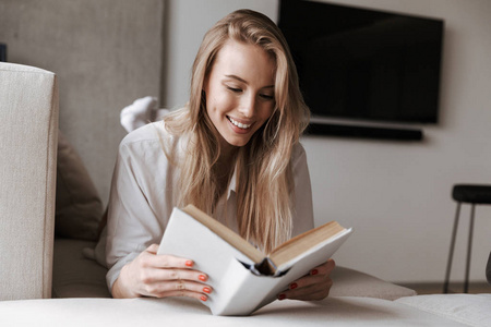 微笑的年轻妇女读空白盖子打开书, 而放松在沙发上在家