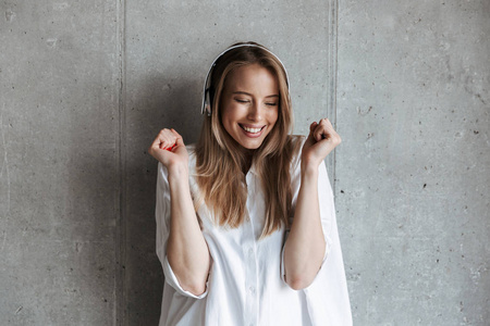 穿着白色衬衫的快乐的年轻女子, 站在灰色的墙壁背景上听音乐与耳机
